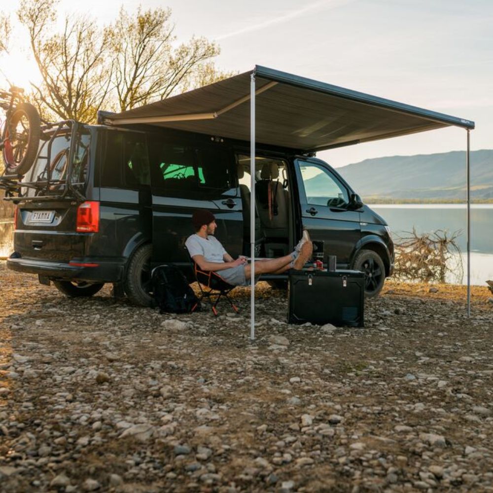 Person sitting by a camper van with an extended awning near a lake.