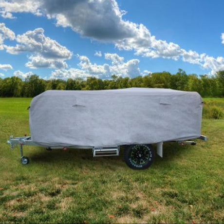 A caravan covered with a gray protective cover, placed on a grassy field with trees and a partly cloudy sky in the background.