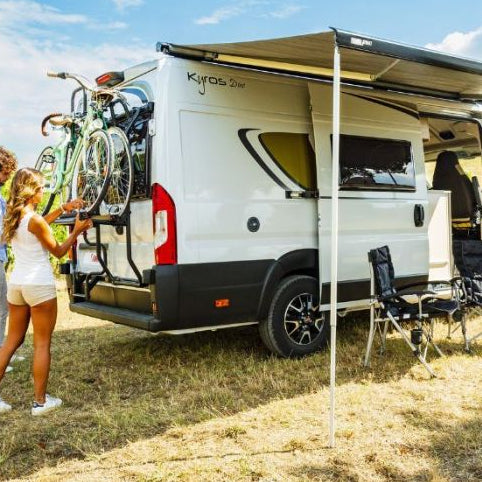 White awning by a vehicle, with FIAMMA branding visible on the side, set up in an outdoor setting with people and chairs nearby.