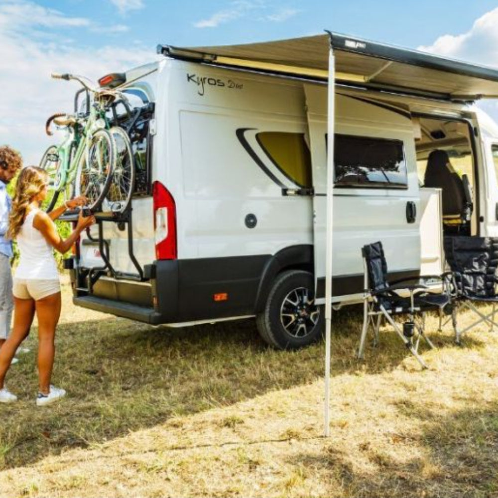 A white awning by a vehicle, with a FIAMMA branding visible on the side, set up in an outdoor setting with people and chairs nearby.