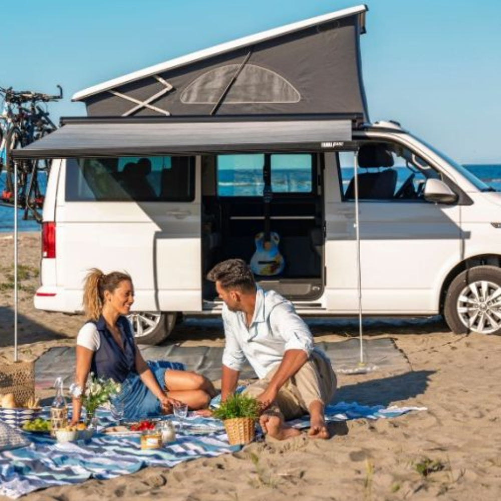 A couple sitting on a picnic blanket by a campervan which has an extended Fiamma F45 S awning providing shade. There are bicycles on the back of the campervan and the ocean in the background.