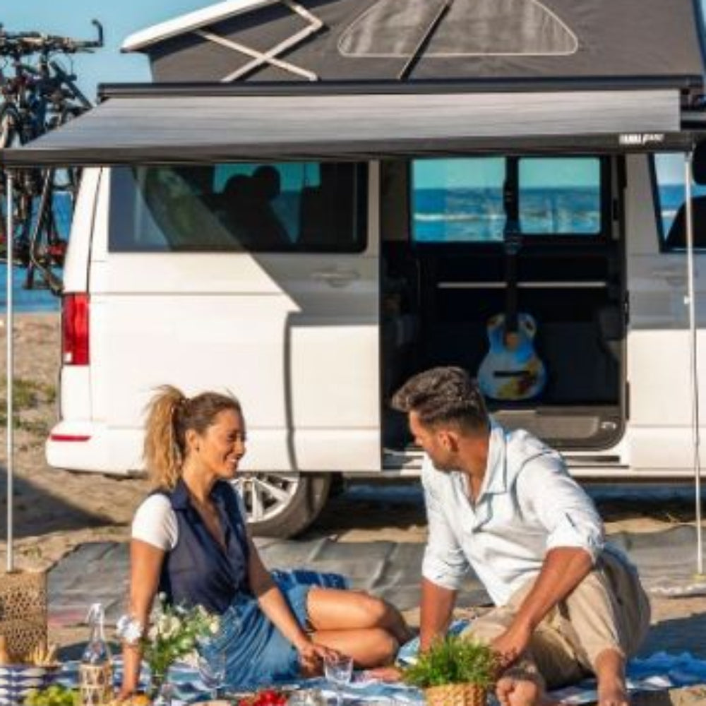 A couple sitting on a picnic blanket by the beach, using a Fiamma F45 S awning attached to a campervan.
