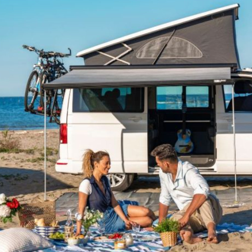 A couple sitting on a picnic blanket by a campervan which has an extended Fiamma F45 S awning in gray. The awning is attached to the side of the campervan and there are bicycles stored on the back of the campervan.