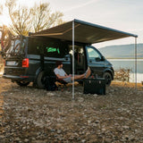 Person sitting by a camper van with an extended awning near a lake.